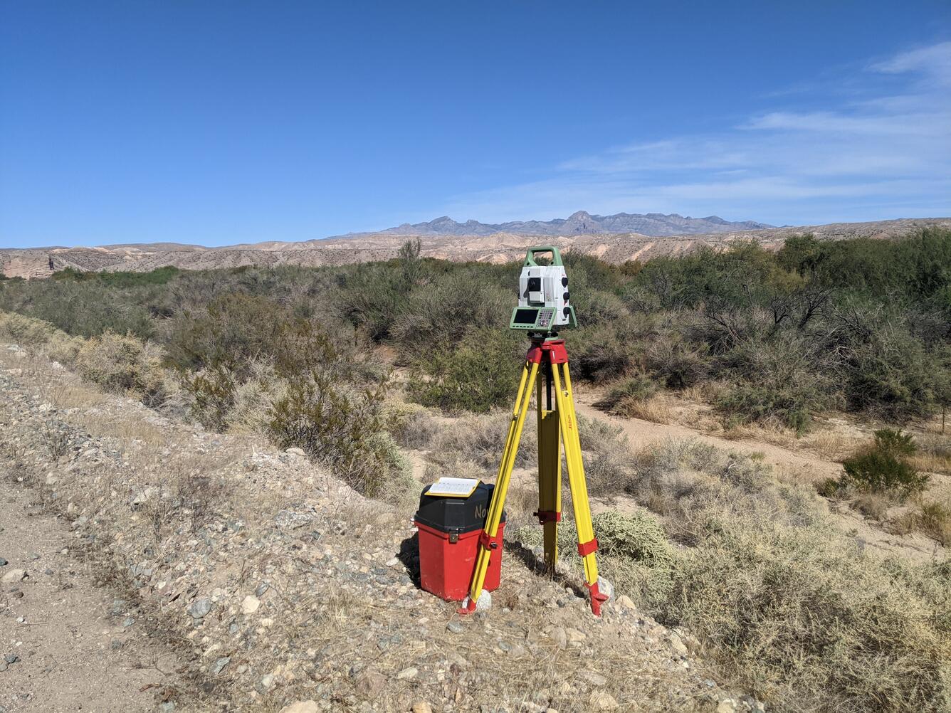 Terrestrial laser scanner on a yellow tripod in a floodplain with brush, distant mountains, and blue sky. 