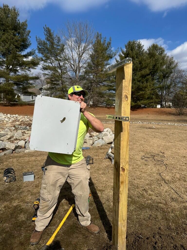 In a field, a man hoists a metal gage box onto a wooden frame.