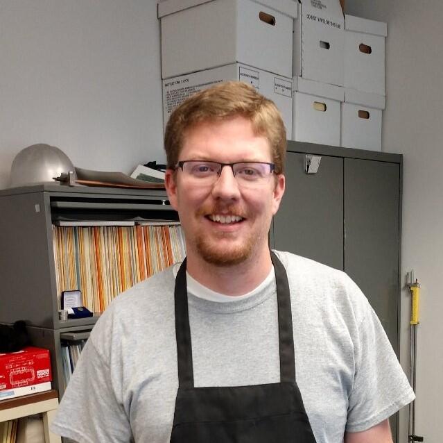 Tom stands in his office in front of metal cabinets with top part of apron showing.