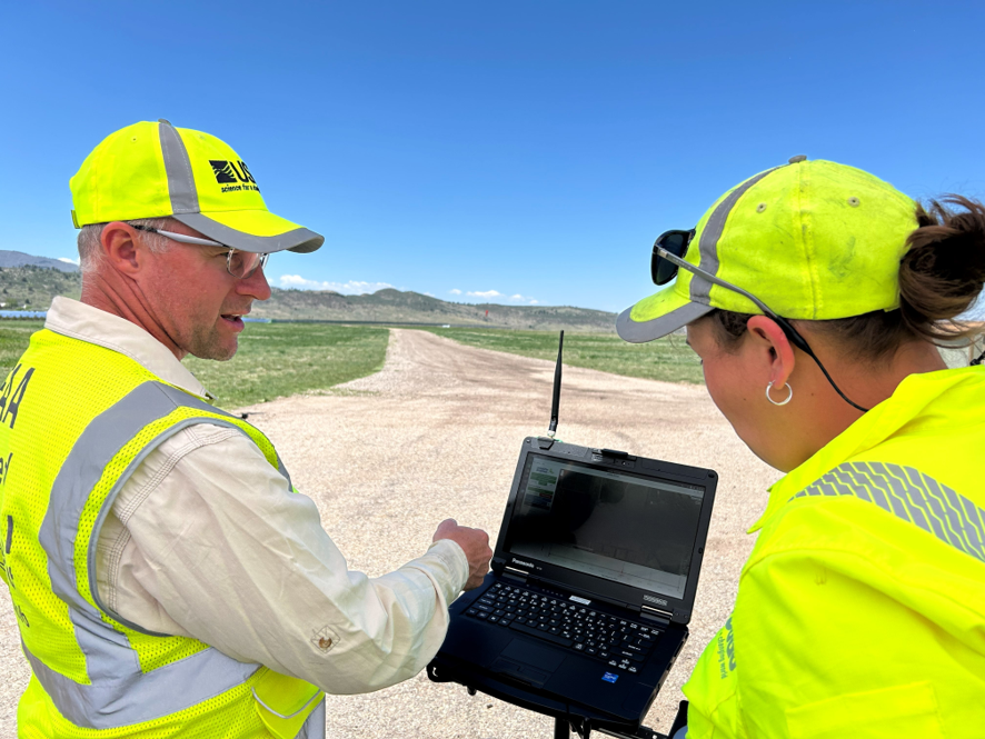 two people in bright yellow USGS gear chat outside while looking at a small computer screen