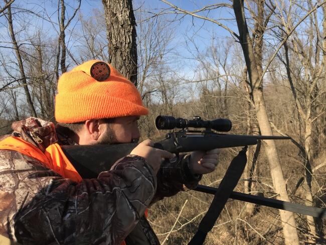  hunter wearing a blaze orange beanie and camouflage jacket aims a bolt-action rifle with a scope.