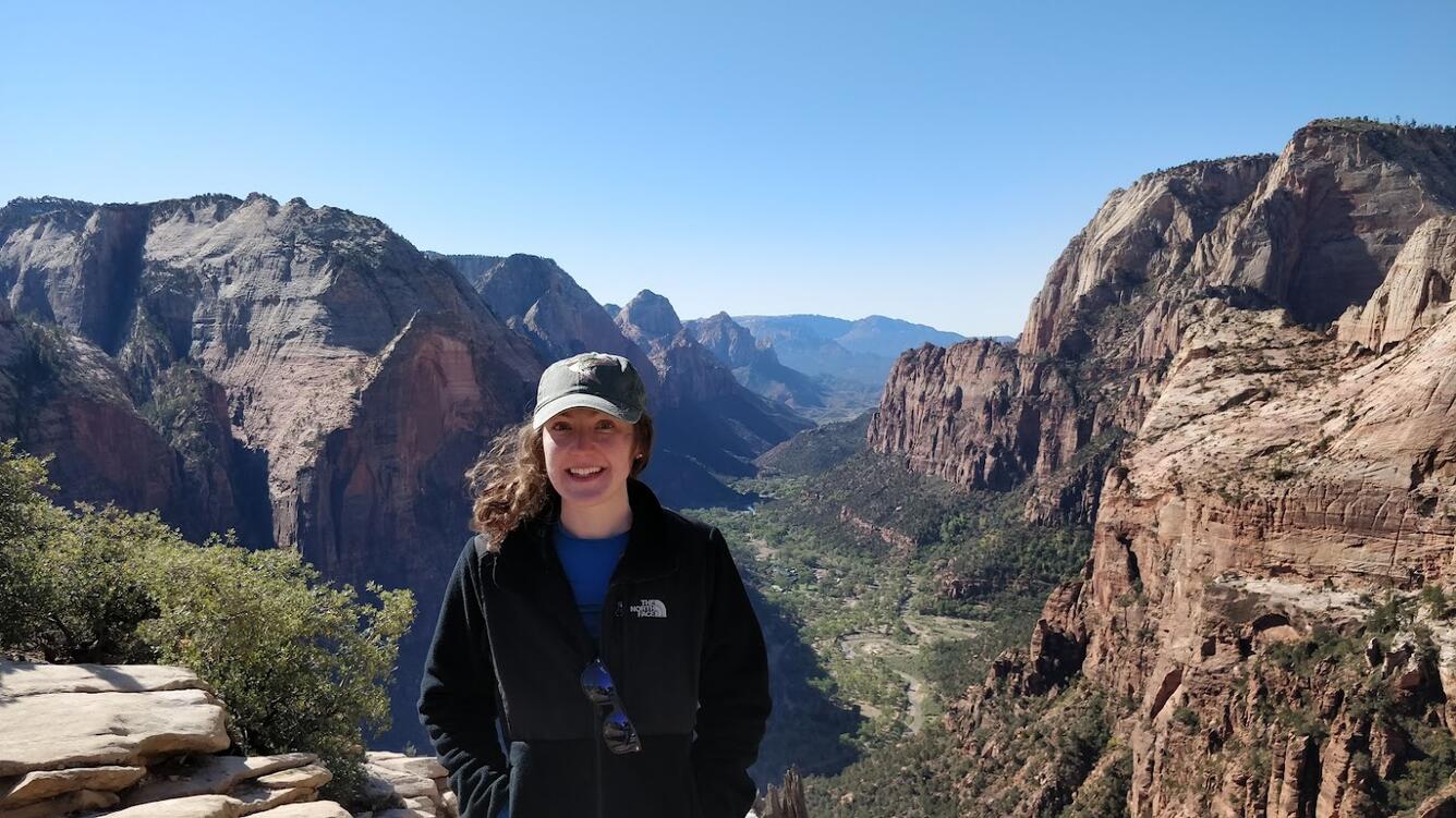 A woman in a black jacket and blue shirt stands in front of a canyon for her picture while wearing a baseball cap over her long, dark hair.