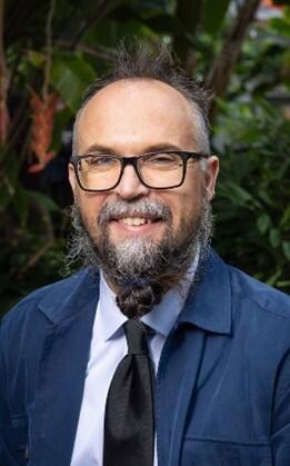 A man with glasses and a beard in a blue jacket, white shirt and tie smiles for his picture.