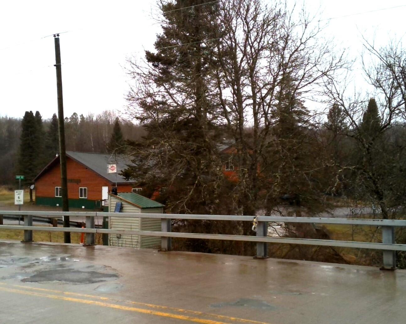 USGS streamgage housing behind bridge guardrails, with a wet road surface on a rural bridge in the foreground.