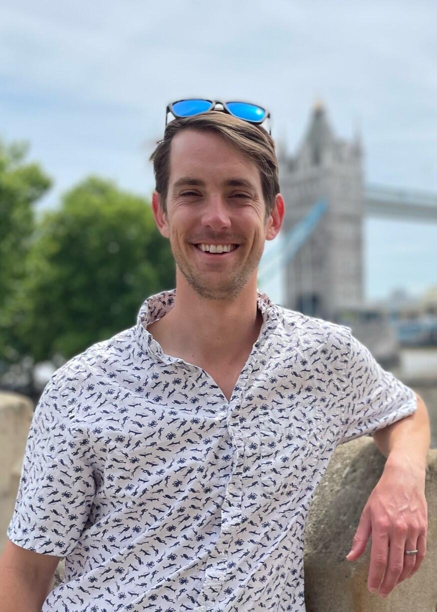 A man in a light colored shirt stands in front of an out-of-focus background on a sunny day.