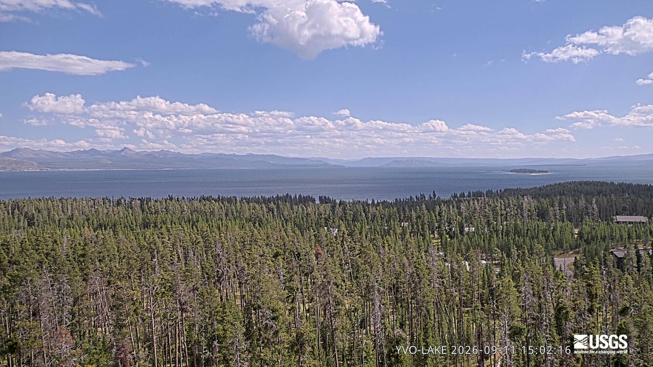 forested area in teh foreground, lake in the middle around, and mountains in the background under partly cloudy sky