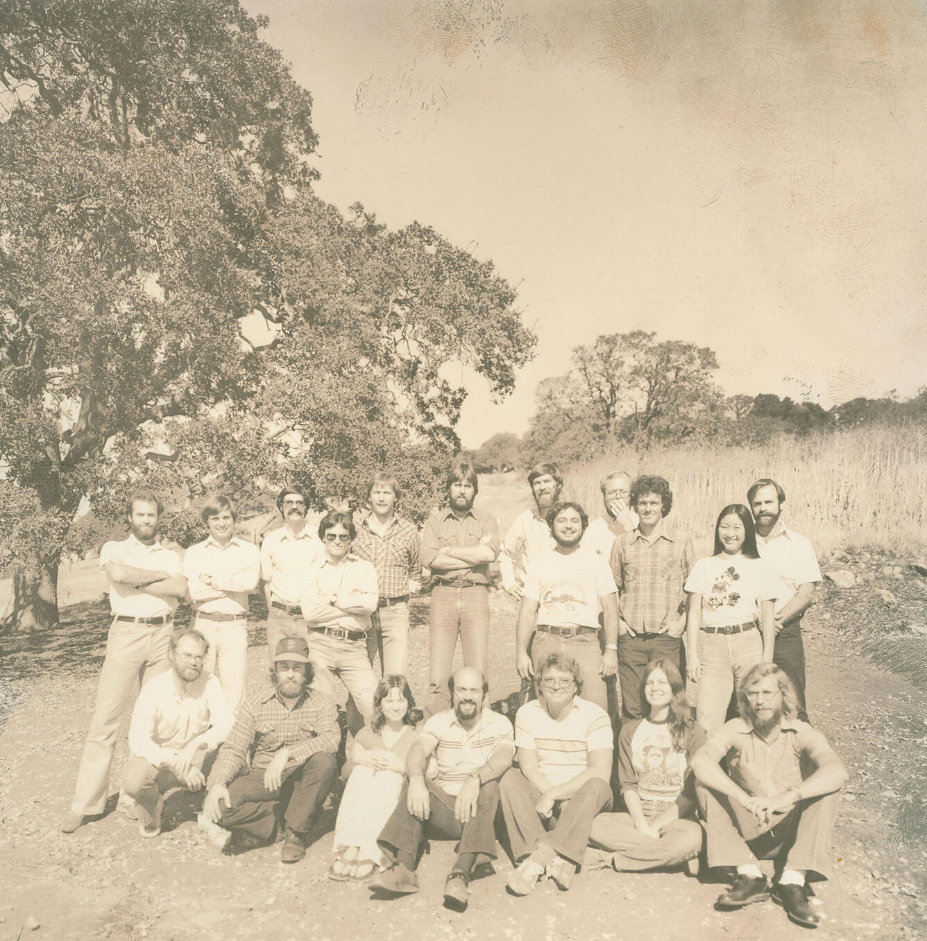 A vintage photo shows a group of nineteen smiling people posing together on a grassy hillside near trees.
