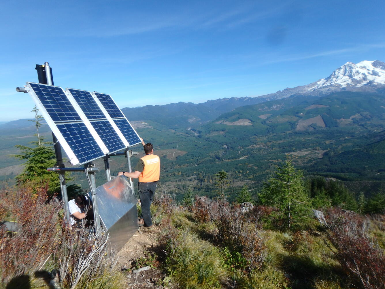 Scientist standing next to solar panels on monitoring station with snowy mountain in the background.