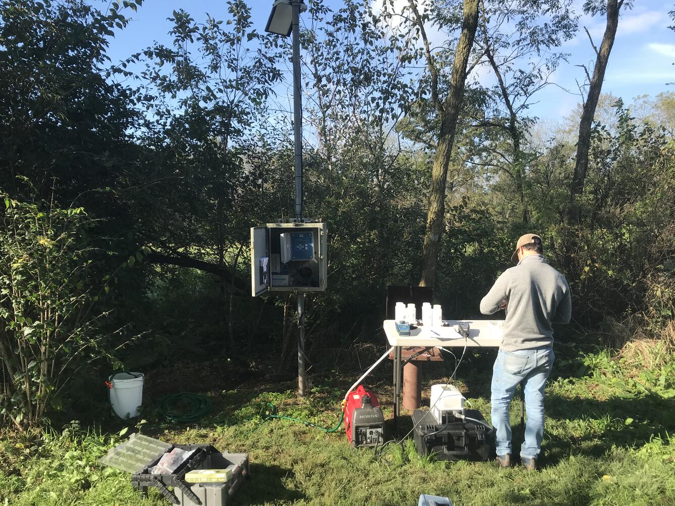 Water-quality sampling at a well located along a narrow tree row. Sample bottles sit on a small table. A scientist records data.