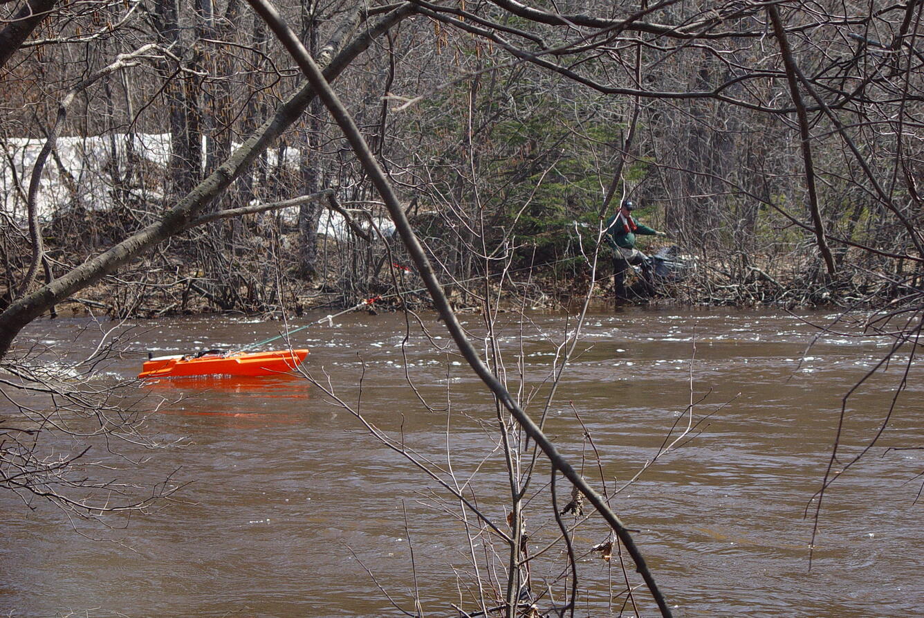 Acoustics boat with ADCP in river with tree branch in foreground, across river is USGS Hydro Tech holding line to boat.