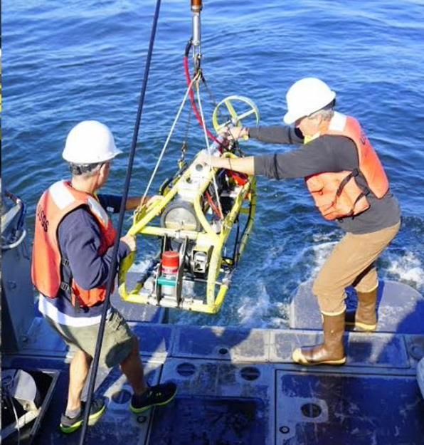 Two men wearing safety gear maneuver a metal frame carrying instruments 