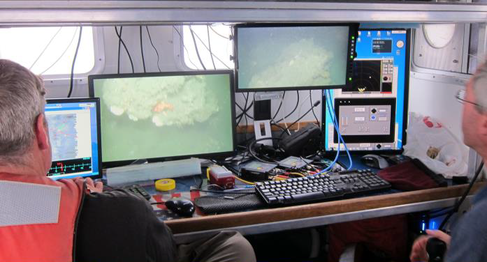 Two men watch computer monitors.