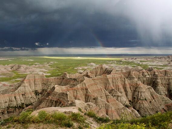 Red-striped tan rock rises, jagged, out of a vast, flat, green valley under very dark clouds of an impending storm.