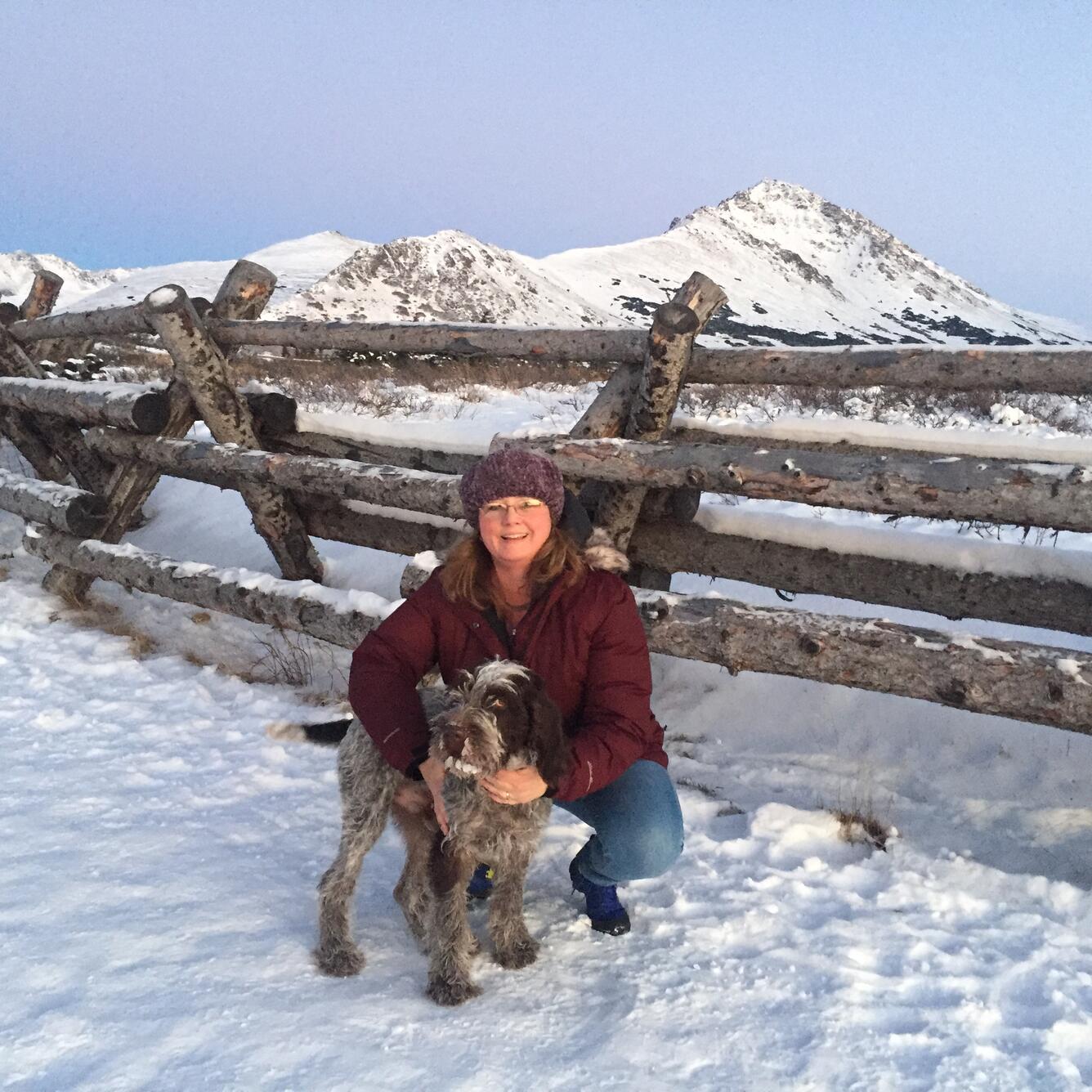 Bobbi Pierson is stooping next to her dog next to a fence in the snow with mountains in the background in the Alaska winter