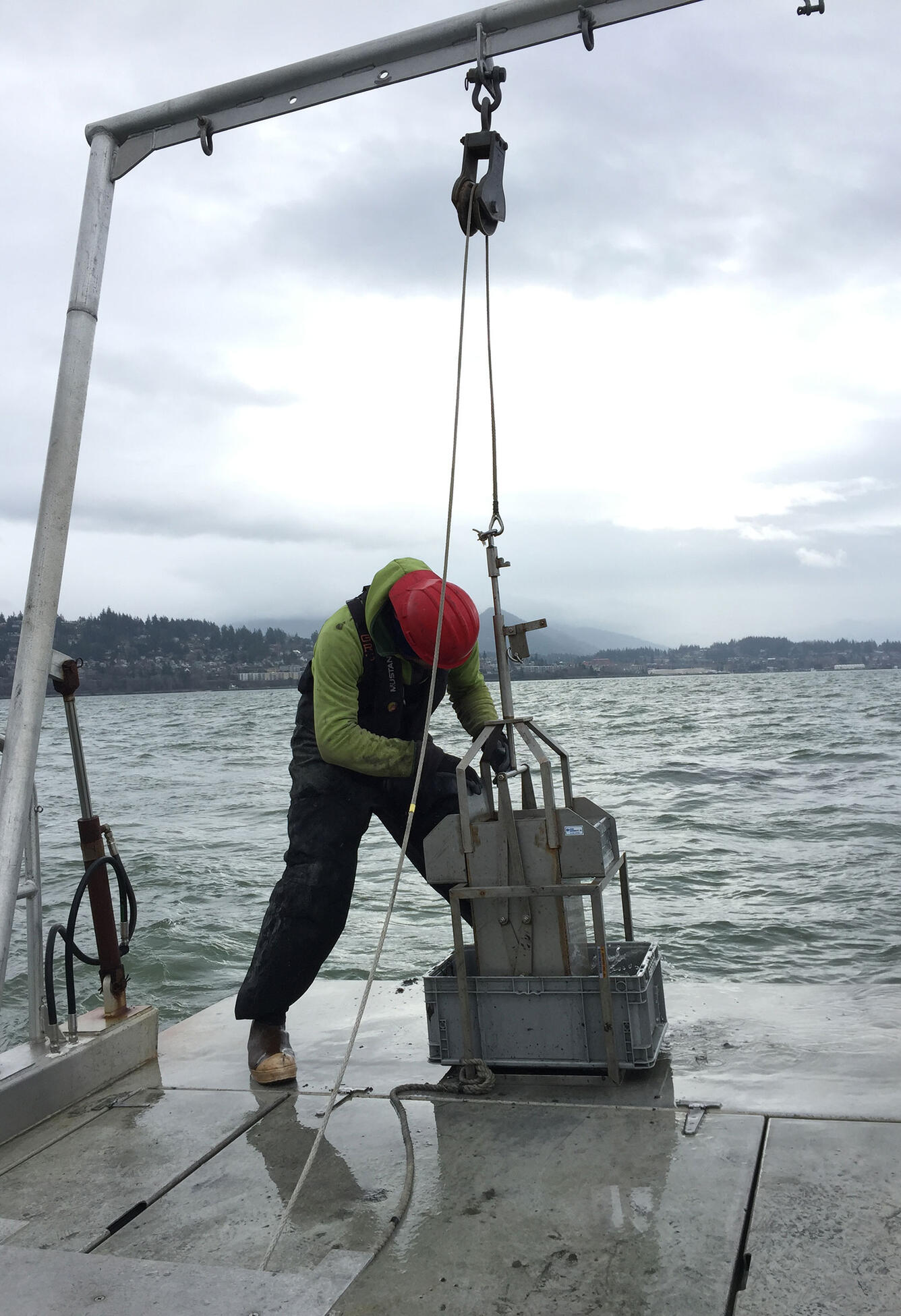A man wearing safety gear stands on the fantail of a small boat preparing a sample-collecting device attached to a pully system.