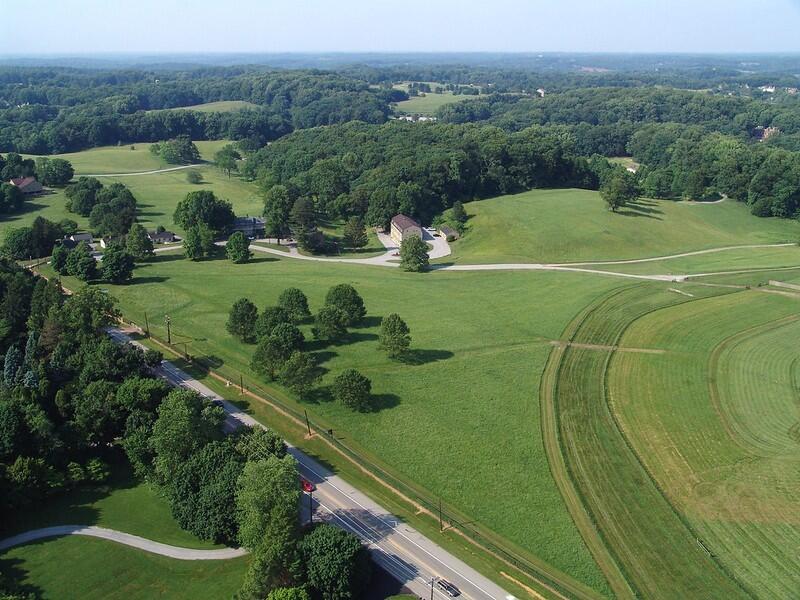 A two-lane highway cuts through agricultural fields interspersed by large clumps of trees.