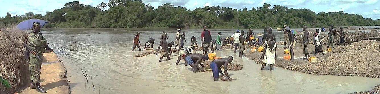 Artisanal miners stand along river while washing gravel, looking for diamonds and gold. A soldier stands guard on shore.