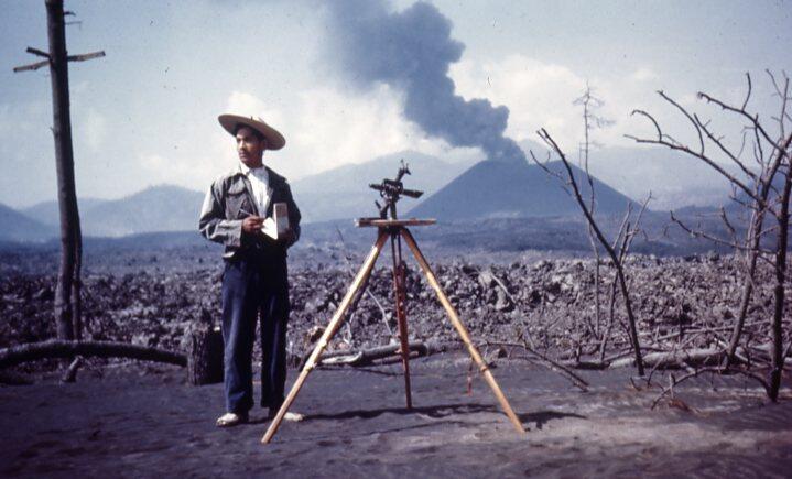Man standing next to surveying equipment with smoky volcano in background
