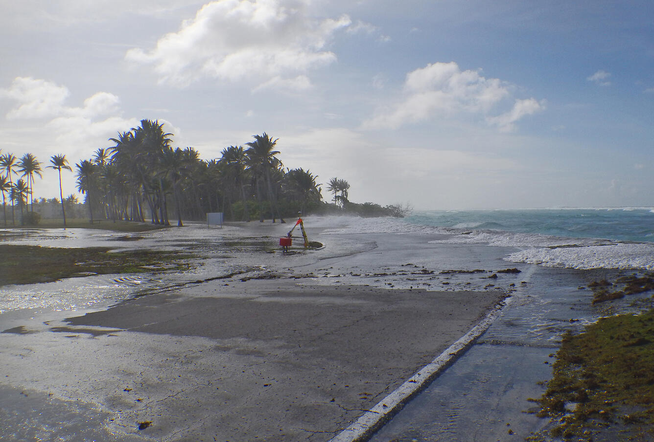 coastal road and palm trees on a small flat island, with water waves from the sea washing over the road