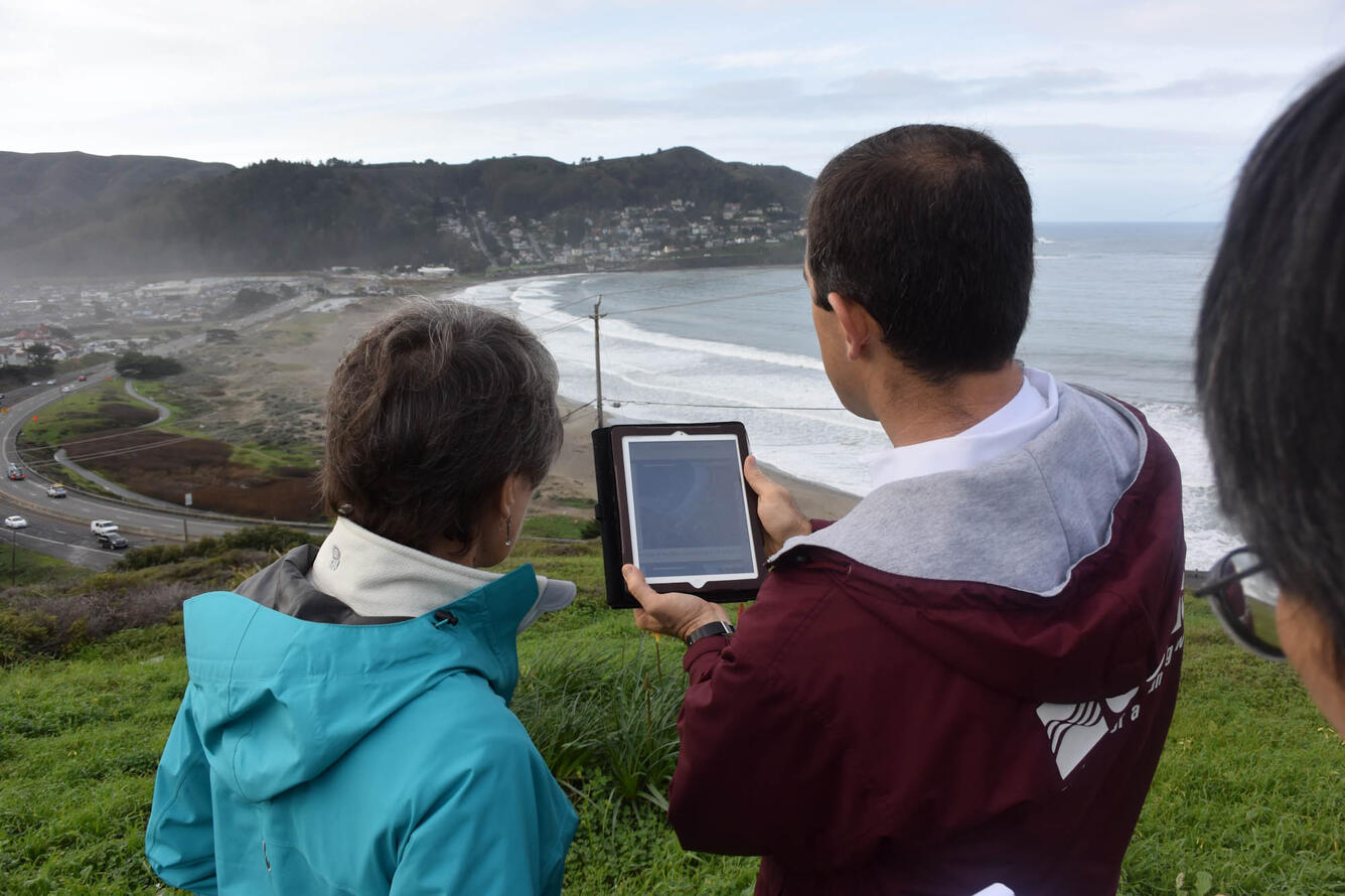 People with backs to the camera on hill above beach, man on right holds an electronic tablet and the woman on the left looks on.