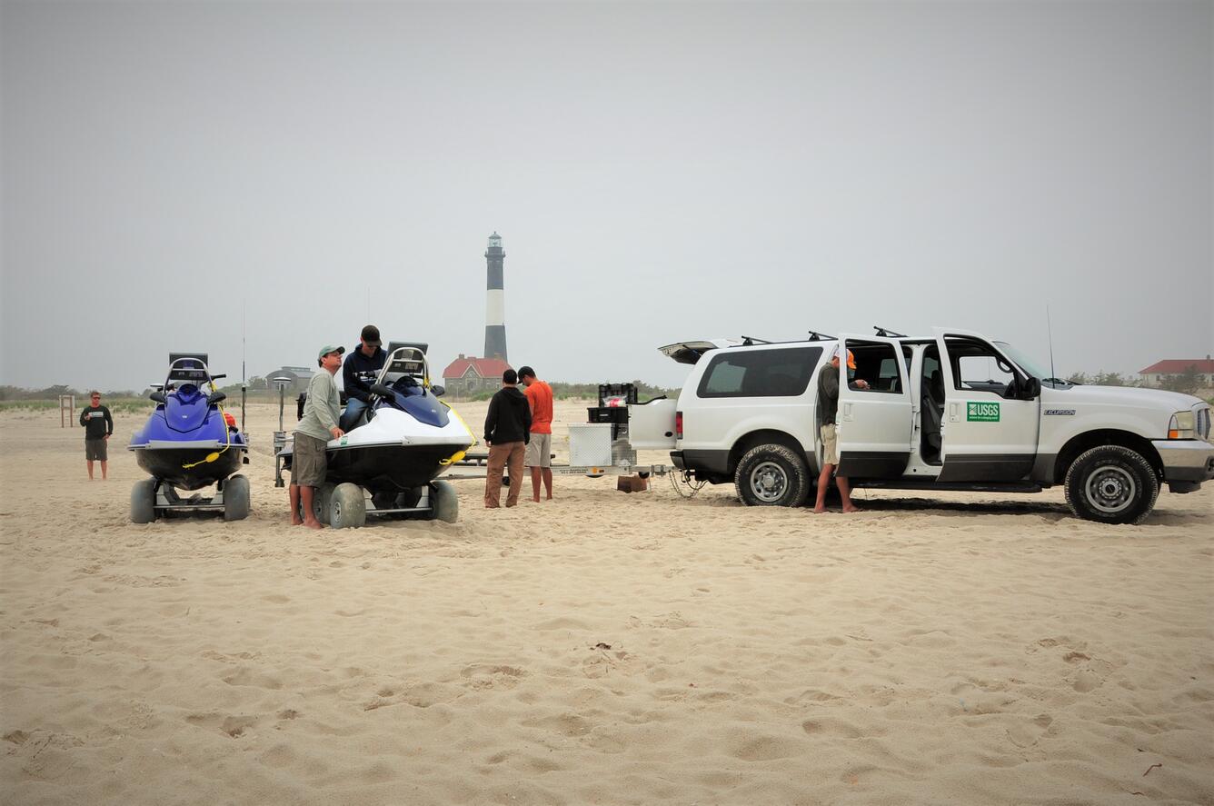 A group of scientists unloads scientific equipment from a truck on a beach. Two personal watercraft sit on carts.