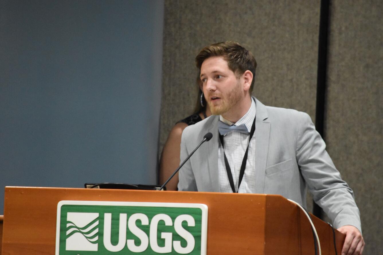 Young man speaks, while standing behind a podium with a USGS sign. A man and a woman stand behind him, watching while he speaks.