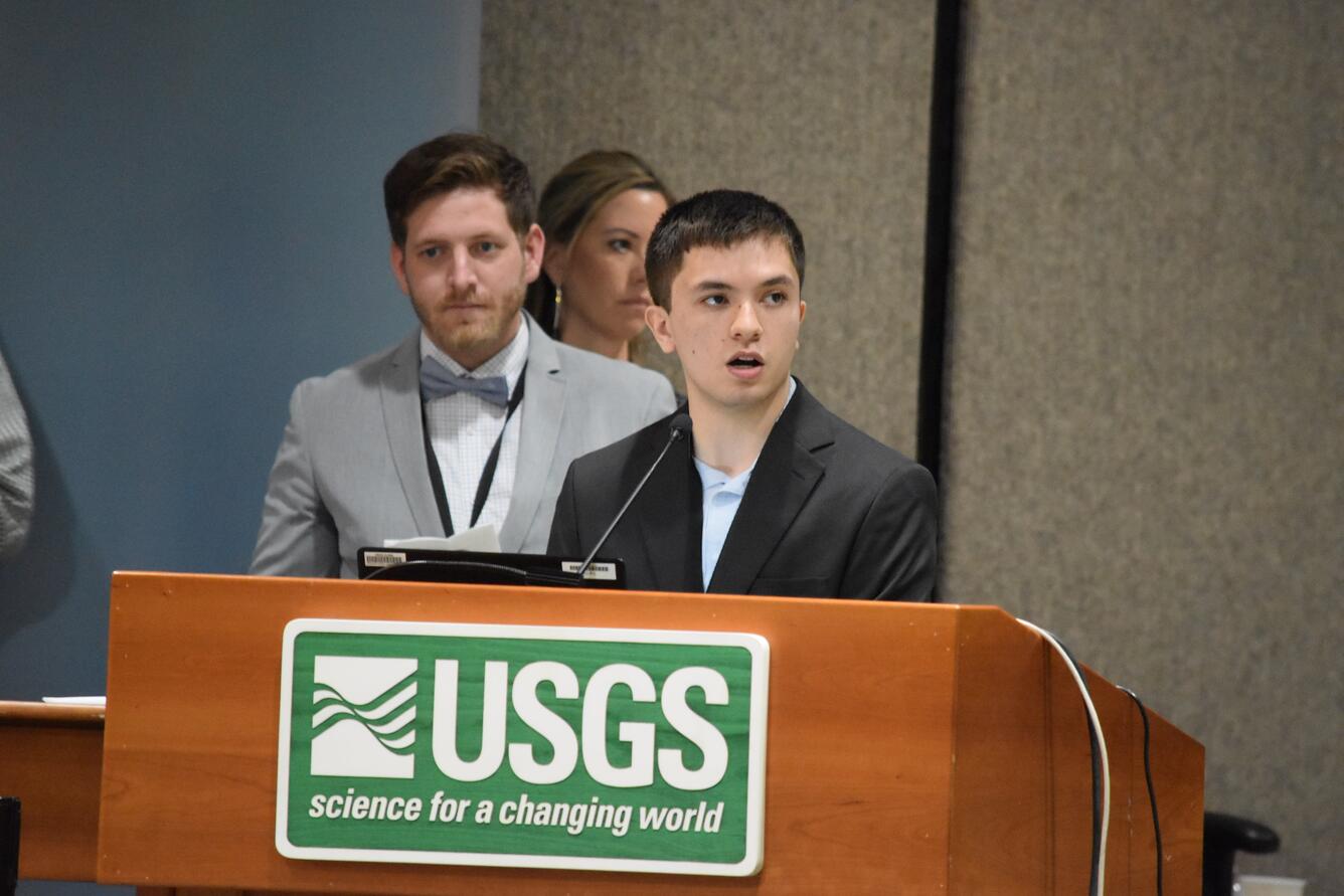 Young man speaks, while standing behind a podium with a USGS sign. 