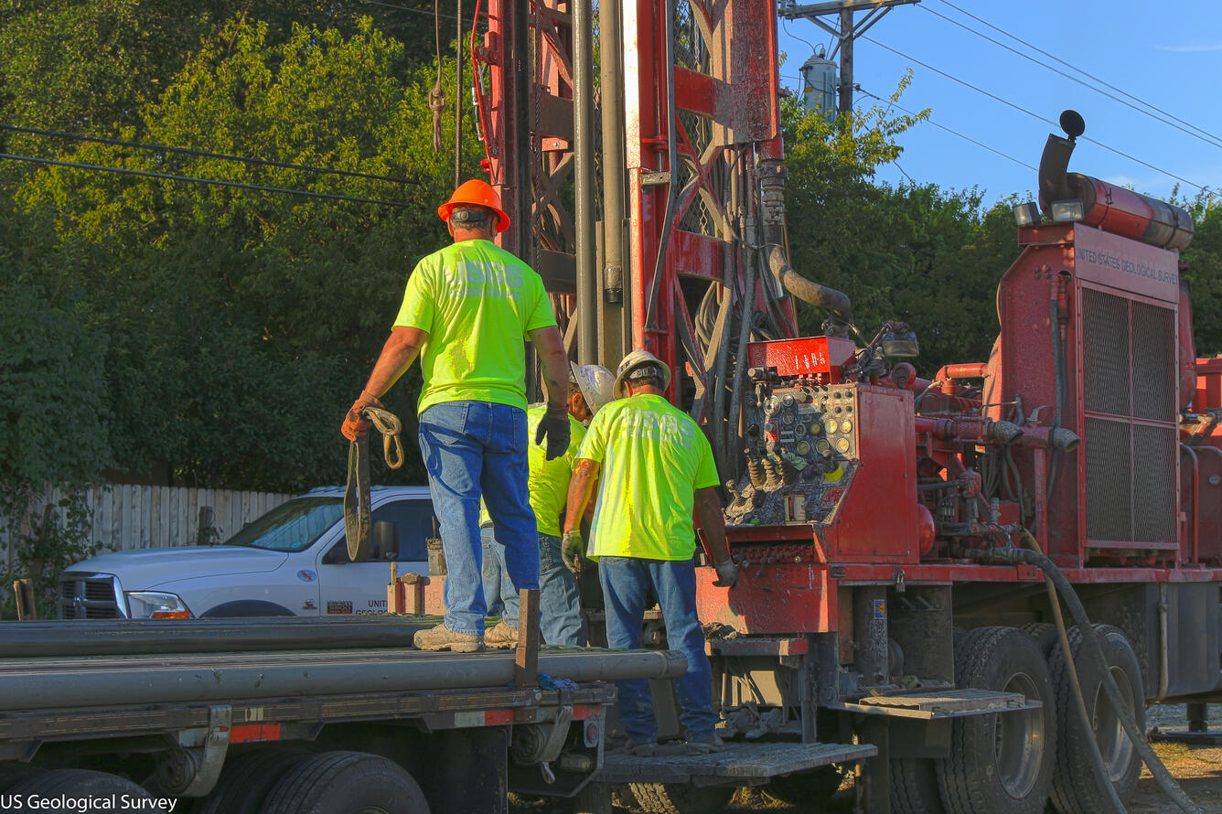 Image shows USGS scientists standing beside a drill rig in protective gear.