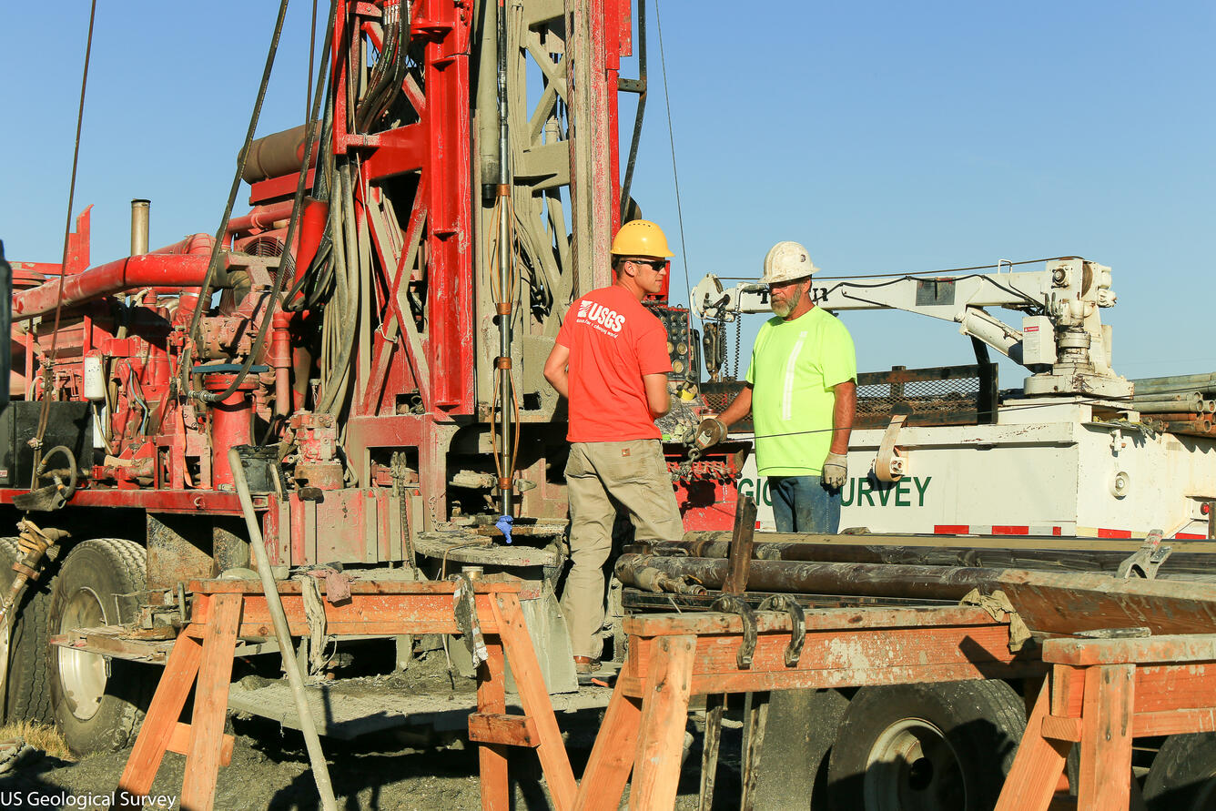 Image shows USGS scientists standing beside a drill rig in protective gear.