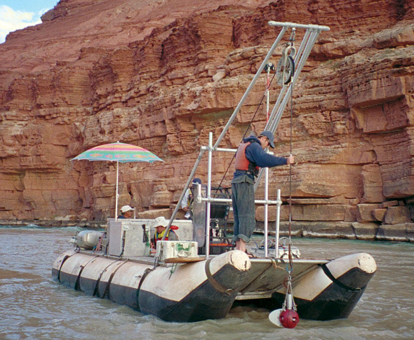 A pontoon boat floats on a river through a red-rock canyon and a man guides an instrument suspended from a cable into the water.