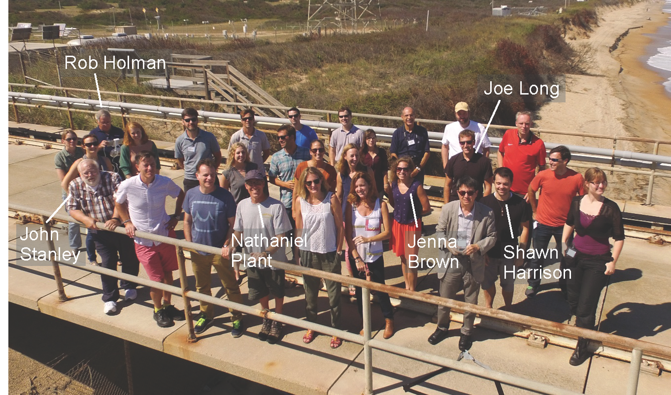 28 people 6 with name labels stand on wooden bridge in bright sunlight. Behind, a beach and grassy bluff stretch into distance