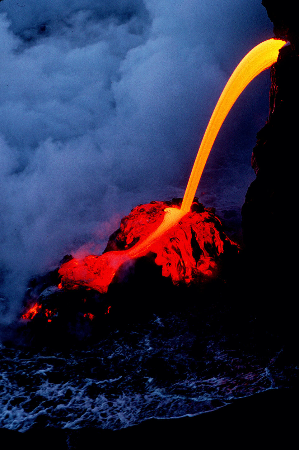 A narrow stream of yellow-hot lava flows out of a lava tube onto rocks and into the ocean.