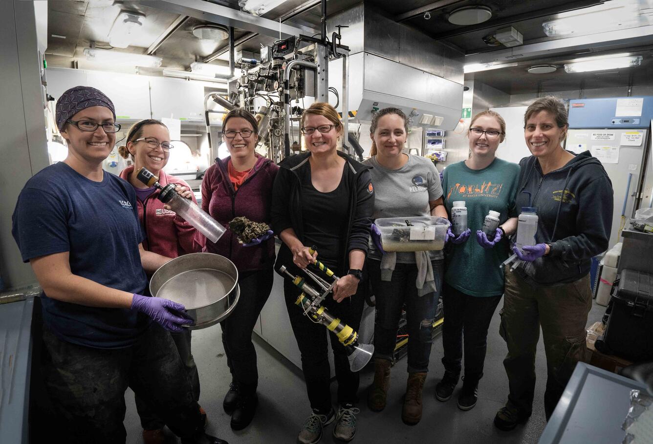 Seven female researchers stand together, smiling, and holding various sediment sampling apparatus.
