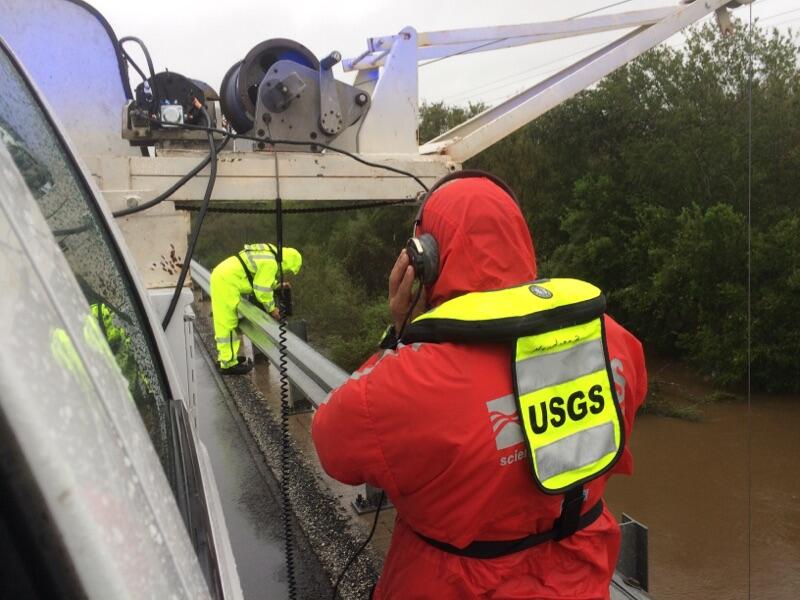 Image shows a USGS scientist in a PFD working with scientific instruments above a flooded river