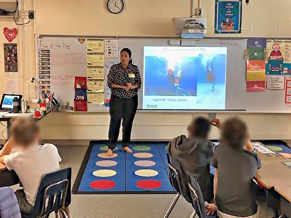 Photo of a woman standing in front of a class of children with an image projected on the whiteboard behind her