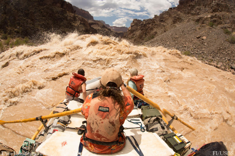 Several people on a raft going through muddy rapids. 