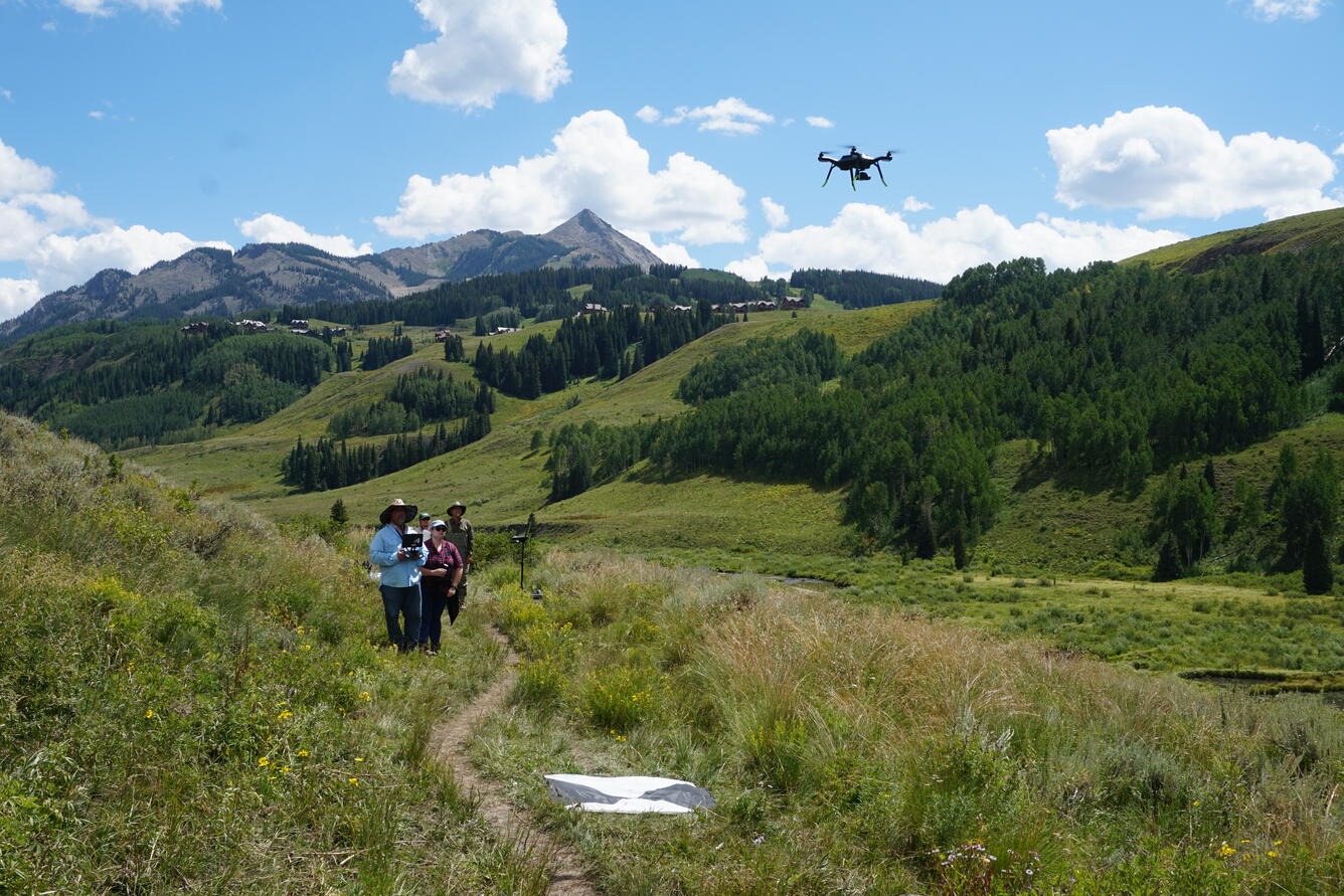 Scientist operates small drone over river valley with mountains in the background.