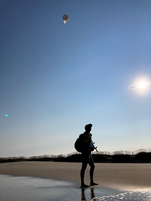 A silhouette of a person on a beach with small white balloon high above against a blue sky