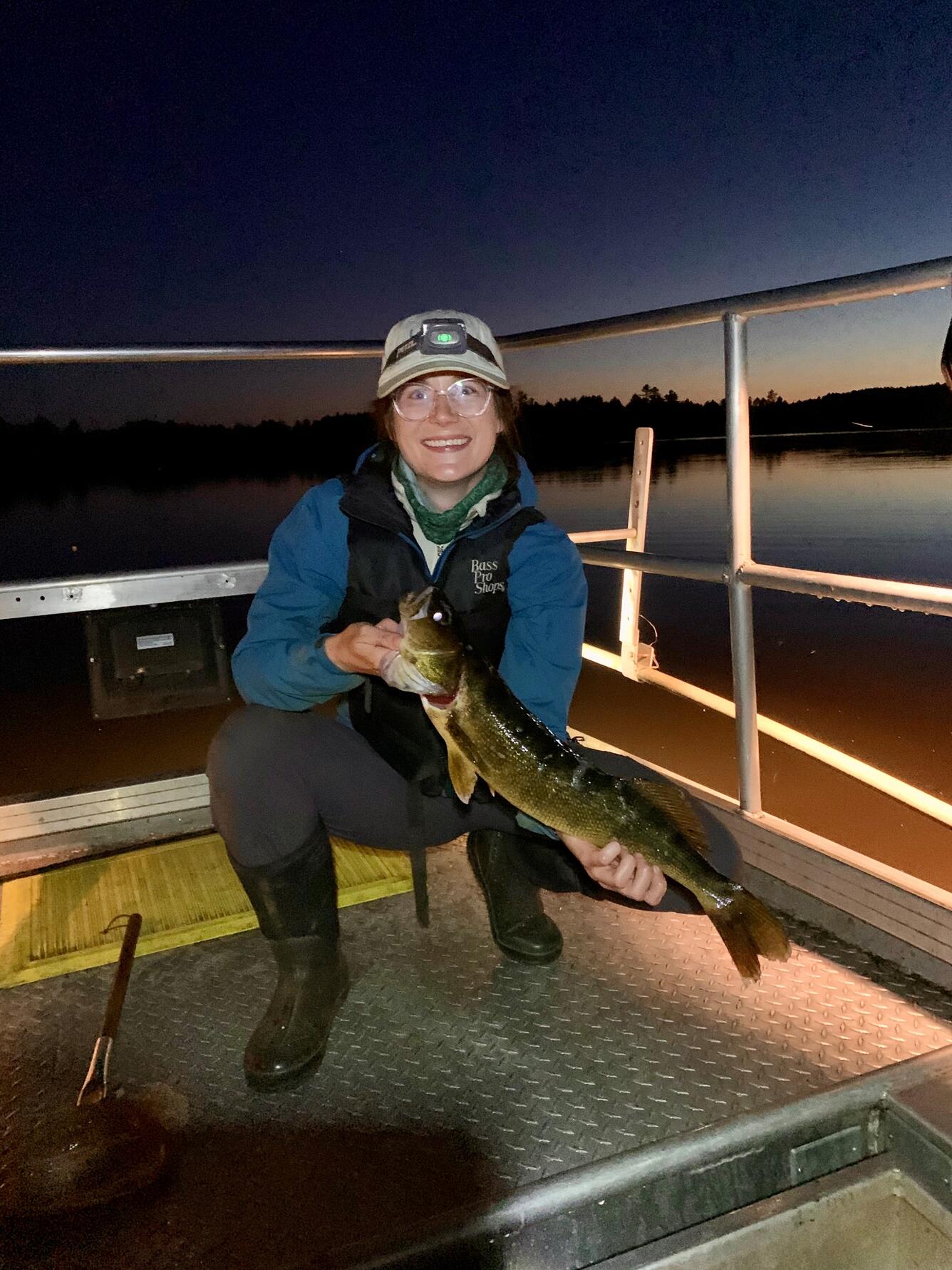 Holly Embke holding a fish with both hands, kneeling & smiling at the camera surrounded by railings on a boat under a dark sky