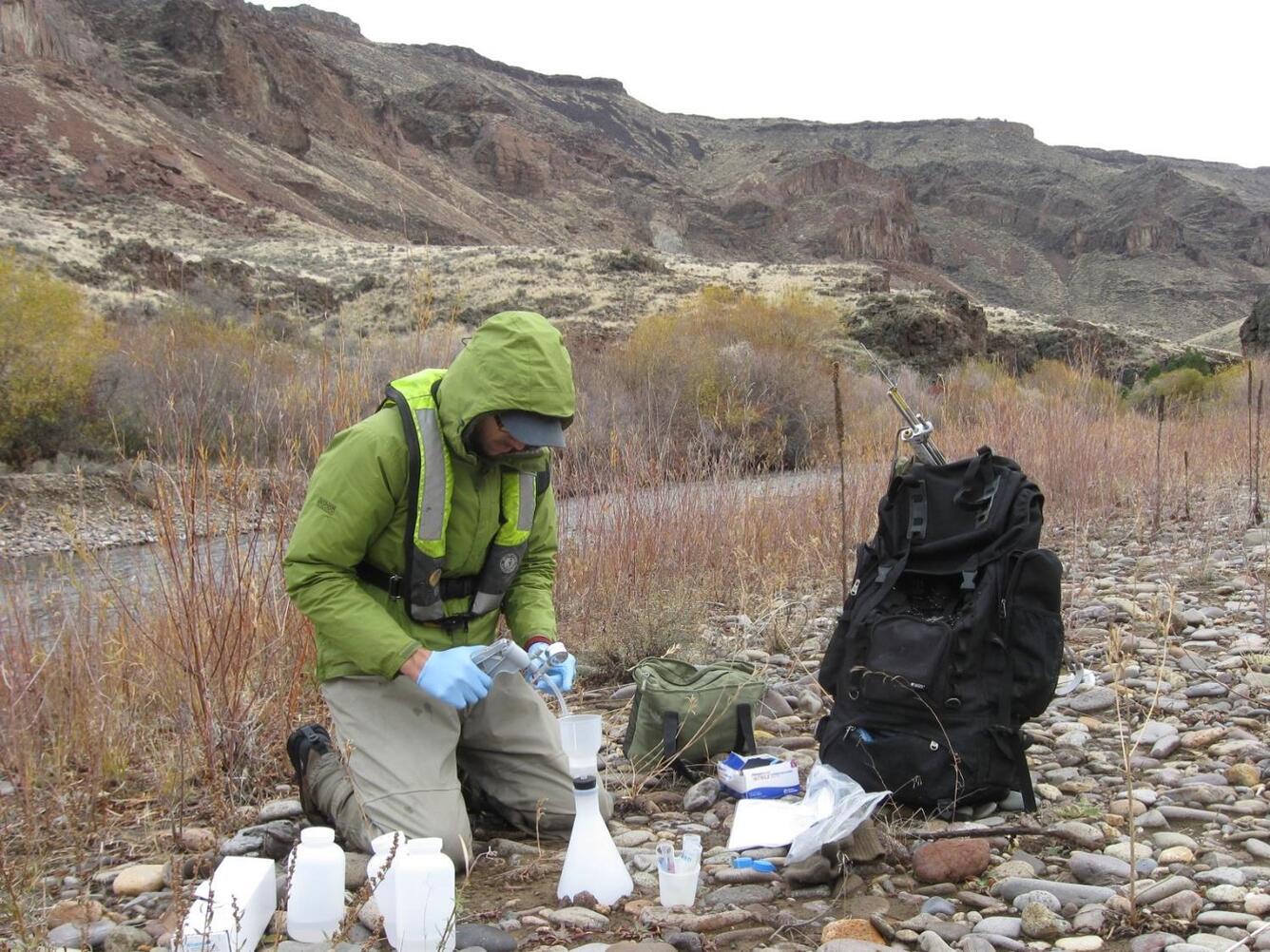 person kneeling on stream bank surrounded by sampling equipment