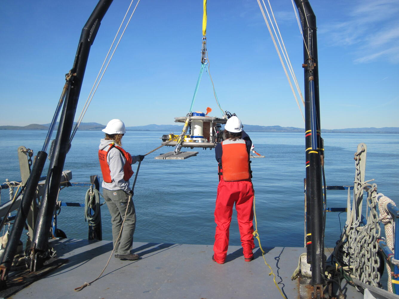 Two women stand on the stern of a boat, they are wearing hard hats, life jackets, steel-toed boots, maneuvering an apparatus.