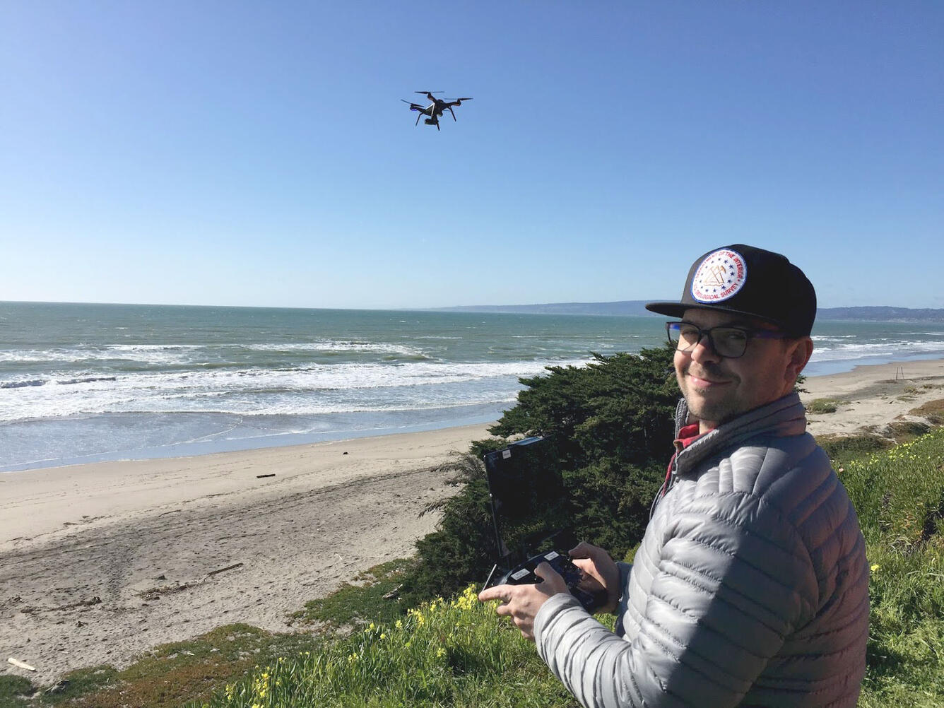 A man stands near the beach in front of a shrub, holding a controller, with a drone flying over the beach behind him