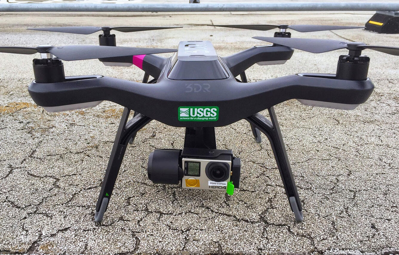 A closeup photo of a black UAS (drone) sitting on the pavement with a USGS sticker on its front and a camera attached