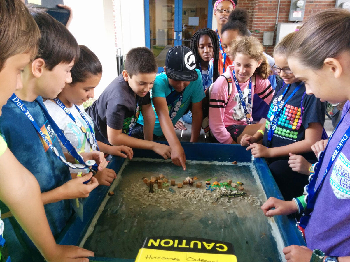 A group of school kids stand around a box with models of structures like houses placed on sand.