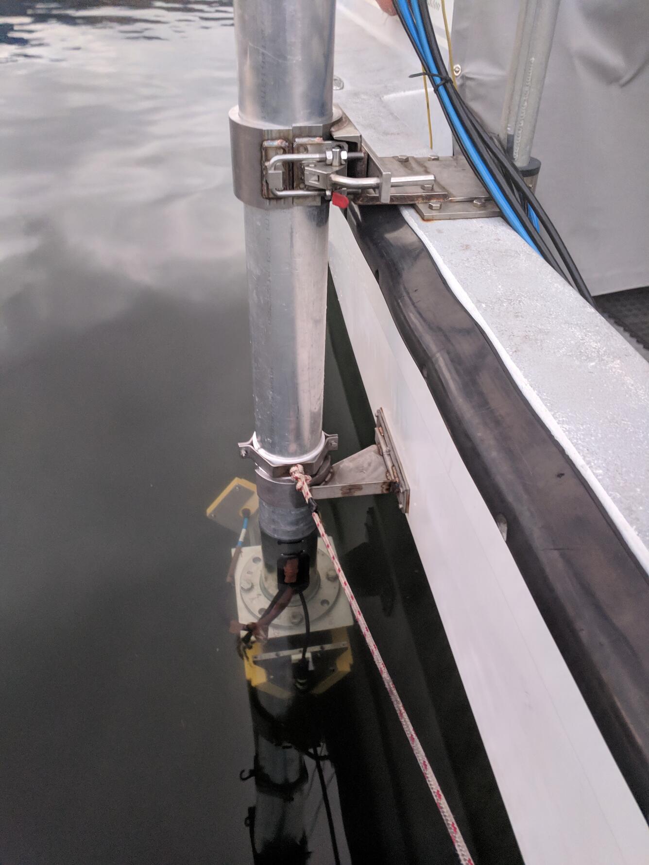 A view of the side of a boat with an instrument attached to a tall, cylindrical pole bolted to it.