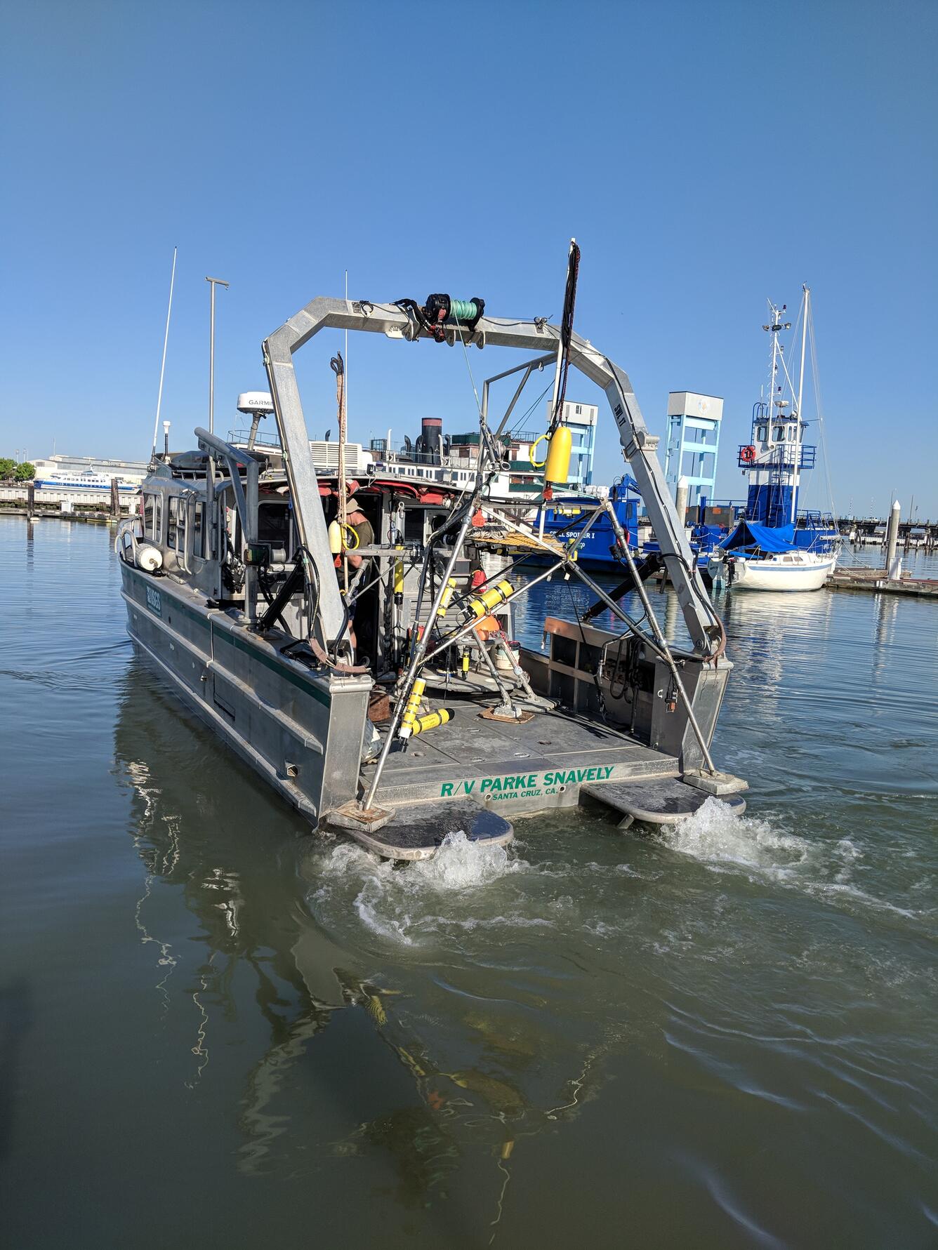 A boat carrying lots of equipment motors out of a yacht harbor, waters are calm, engines churn up the water at the stern.