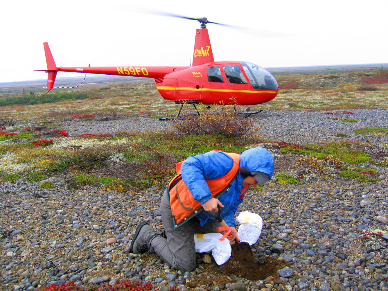 Scientist with a helicopter in the background collecting a sample of glacial till, Dillingham quadrangle, AK.