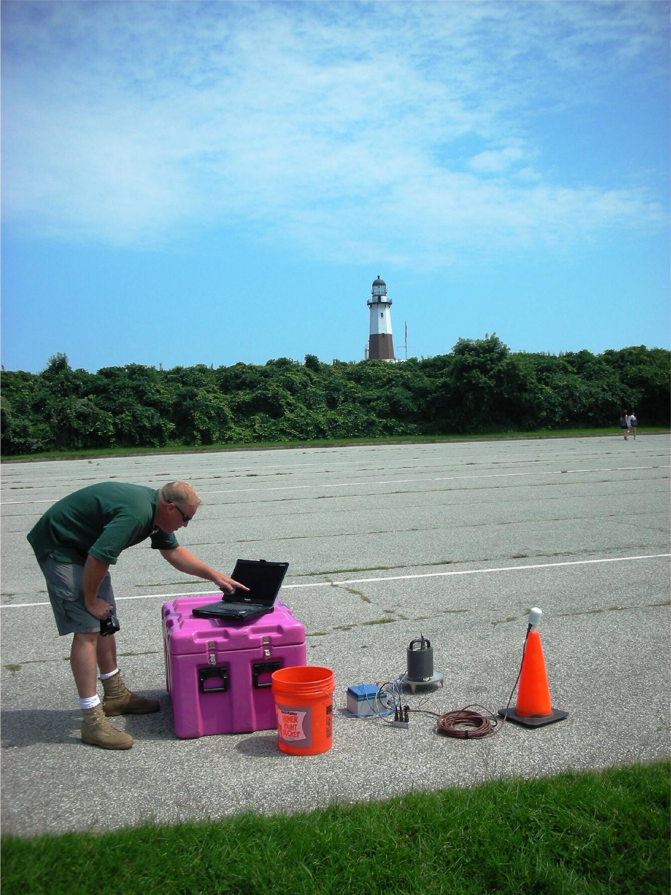 man in green shirt working on laptop on top of pink case on blacktop with lighthouse in background 