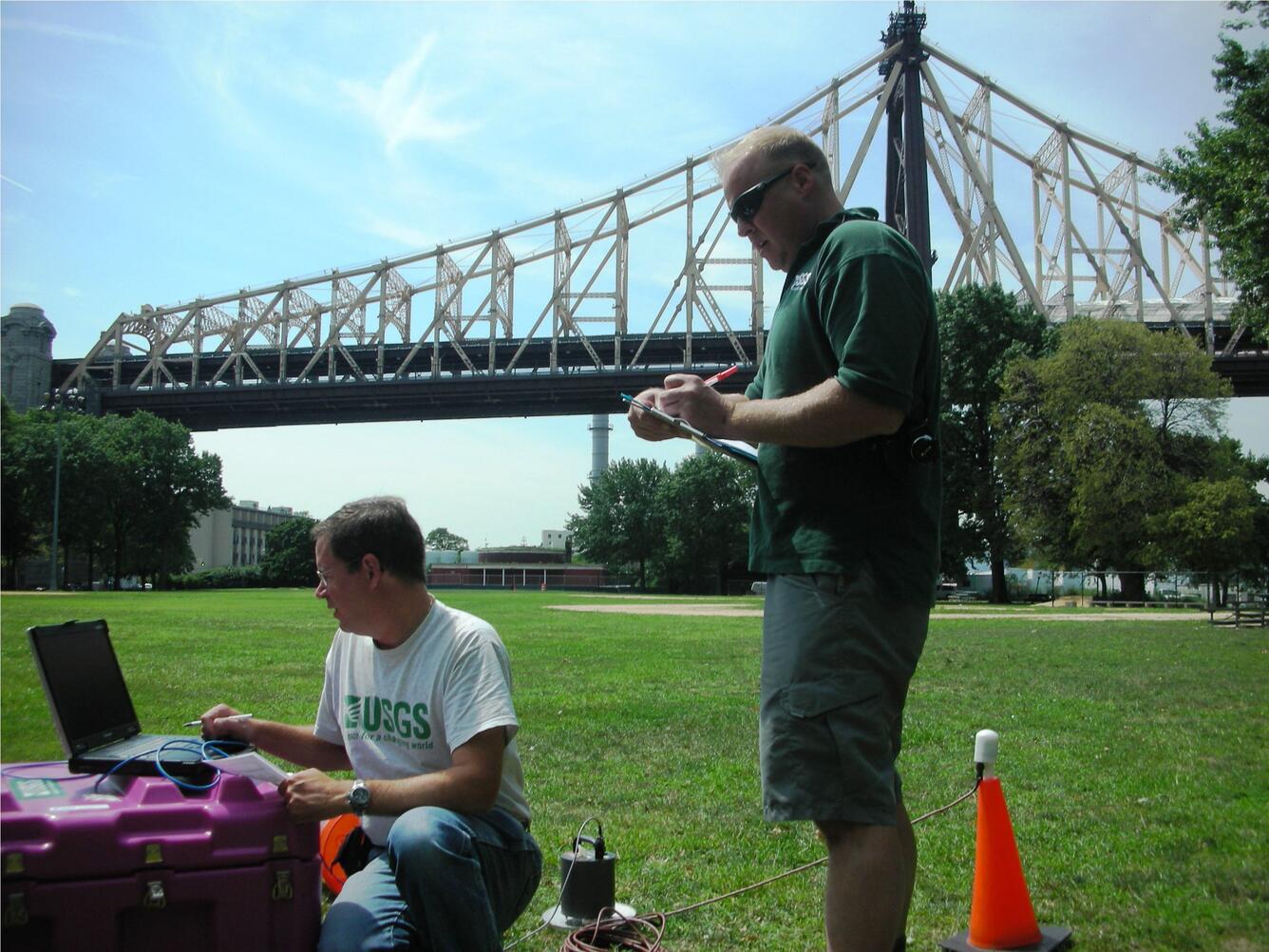 man standing recording data while another man kneeling is working on laptop outdoors with bridge in background