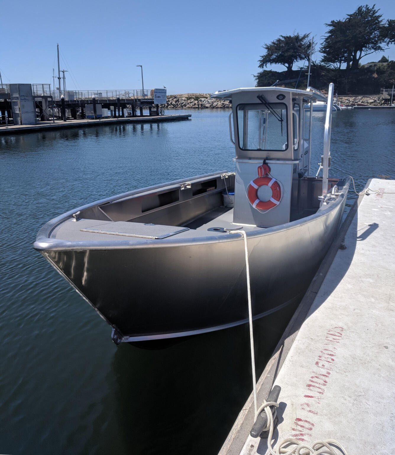 A small aluminum boat with a wheelhouse sits in the water tied up to a dock in a calm boat harbor.